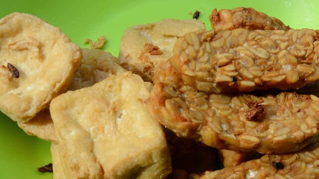 Extreme Close-up of Crispy Fried Tofu (Tahu) and Tempeh Served on a Bright Green Plate. Traditional Indonesian Vegan Food, Plant-Based Protein, and Street Food