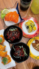 Assortment of sushi and fresh salmon sashimi on wooden table. Two small bowls of spicy squid, restaurant plates, and a green beverage are present
