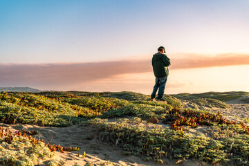 Person standing on coastal dunes at sunset, overlooking the sea in warm golden light.