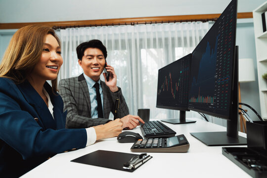 Smiling Asian businessman calling to broker dynamic stock exchange database with woman business partner pointing to screens comparing profit value online application program at home office. Infobahn.
