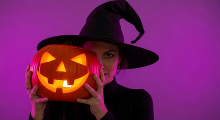 Woman in witch hat holding a glowing jack o lantern against a purple background for halloween night
