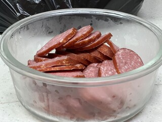 Sliced smoked sausage in a glass bowl, ready for cooking or serving. Close-up food photography on a light kitchen background.