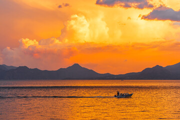 Fiery sunset over Lake Atitlan with glowing clouds above the volcanic mountain silhouette and a small boat crossing the shimmering golden water peaceful dramatic evening views of Guatemala’s iconic