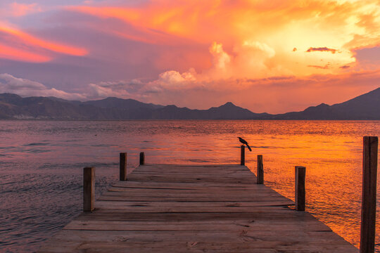 Wooden Pier Extending into Lake Atitlan at Dramatic Sunset with Golden Water Reflections Distant Volcanic Mountains Peaceful Horizon and Solitary Bird Creating Serene Guatemalan Landscape