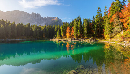 Crystal-clear turquoise water of a high-altitude lake reflecting a pine forest with autumn colors and the majestic, rugged mountains of the Dolomites.