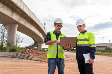 Two Asian civil engineers review plans on a laptop at a highway construction site under a concrete overpass against a cloudy sky.