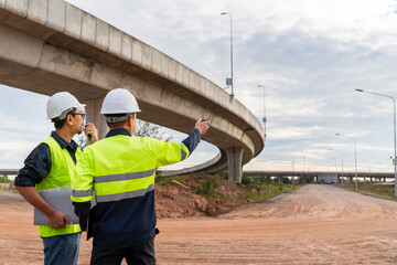 Two construction managers, communicating via radio, inspect a dusty highway site under a curving concrete overpass.