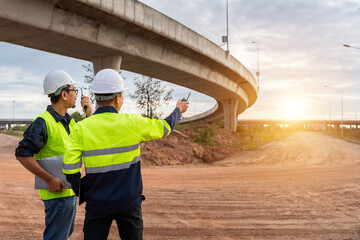 Construction engineers communicating via radio at sunset on a dusty, unfinished highway site under a curved concrete overpass.