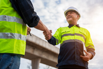 Two construction engineers in safety gear shake hands, finalizing a deal or showing successful teamwork at a construction site.