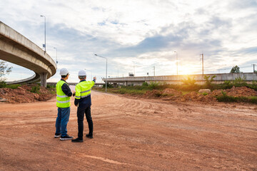 Two construction engineers stand on a muddy dirt road, pointing toward the sunset near a highway overpass on the job site.