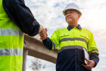 A confident construction manager in a hard hat and safety jacket shakes hands with a colleague at a sunny highway worksite.