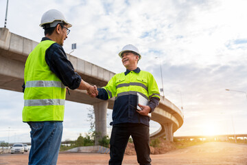 Two Asian engineers shaking hands on a dusty construction site near a curved overpass during a bright sunset.