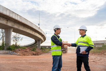 Two construction engineers shake hands on a dusty job site with a dump truck and a curved concrete overpass in the background.