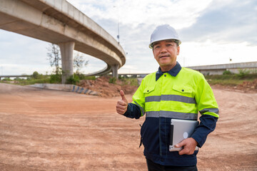 A confident Asian construction manager holds a tablet and gives a thumbs-up on a dusty highway site under a concrete overpass.