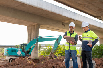 Two construction engineers review plans on a laptop with an excavator and a concrete overpass in the background of the worksite.