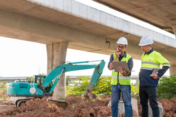 Two construction supervisors using a radio and tablet to discuss plans at a highway worksite, with an excavator under an overpass.