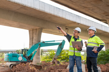 Asian Two construction engineers use a radio and laptop to direct work near an excavator under a towering concrete highway overpass.