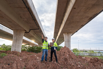 Two construction engineers stand on a high dirt pile, pointing and inspecting the structure of massive dual highway overpasses.