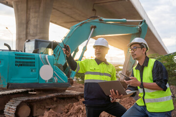 Construction engineers communicating via radio and laptop, directing work in front of a large excavator under an overpass at sunset.