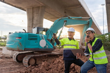 Construction engineers direct operations using a radio and laptop in front of a heavy excavator under a massive overpass.