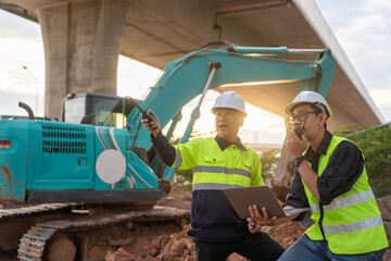 Construction supervisors use radios and a laptop to coordinate work in front of a blue excavator under a concrete overpass.
