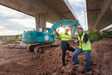Construction engineers stand on a dirt pile, inspecting an excavator and using a laptop under massive dual overpasses.