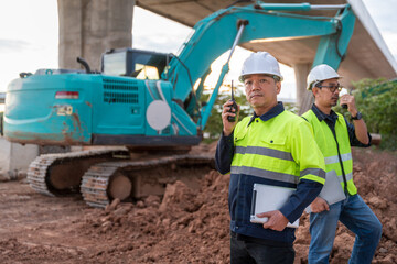 Two construction supervisors, holding tablets and communicating via radio, stand in front of a large excavator at the worksite.