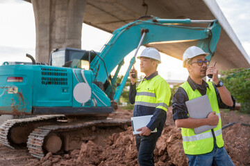 Two construction engineers use two-way radios to coordinate work in front of an excavator under a concrete highway overpass.