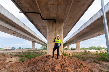 A construction manager stands on a dirt mound, communicating via radio under a massive, symmetrical highway overpass structure.