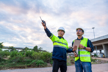 Asian two construction engineers use a two-way radio and tablet to direct operations under a vast, cloudy sky at a highway worksite.