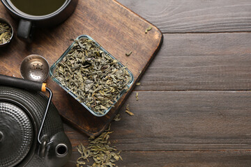 Glass bowl with dried green tea leaves and teapot on wooden background