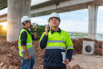 Construction workers in safety gear communicate on site under a bridge during daylight, showcasing...