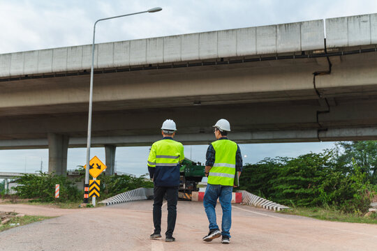 Construction Workers Inspecting Bridge Infrastructure on a Roadway Under an Elevated Highway in Daylight with Safety Gear and Traffic Signs