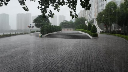 30 October 2025 – Heavy rain falls over a city park in Hanoi, Vietnam. Wet plaza, lake, and high-rise buildings with passing traffic.