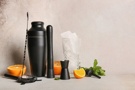 Glass with ice cubes, bartender utensils, orange and mint leaves on beige grunge background