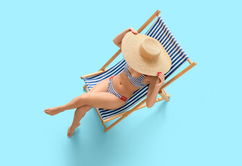 Beautiful young woman in striped swimsuit sitting on deckchair against blue background, top view
