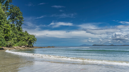 The waves of the turquoise ocean roll towards the shore, foam, and spread out on the sandy beach. No people. Mountains on the horizon. A green hill against a blue sky, clouds. Malaysia. Borneo. 
