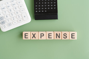 EXPENSE text on wooden cube blocks with black calendar and calculator on green background, top view