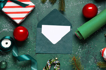 Obraz premium Blank greeting card in envelope with Christmas decorations, gift box and packing materials on grunge green background