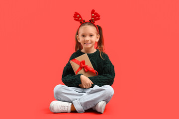 Naklejka premium Cute little girl in Christmas reindeer horns with gift book sitting on red background