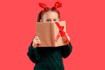 Naklejka premium Cute little girl in Christmas reindeer horns with gift book on red background