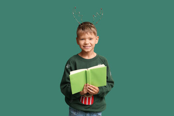 Naklejka premium Cute little boy in Christmas reindeer horns with book on green background