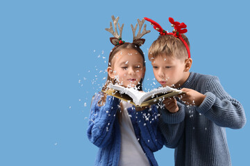Naklejka premium Cute little children in Christmas reindeer horns blowing snow off book on blue background