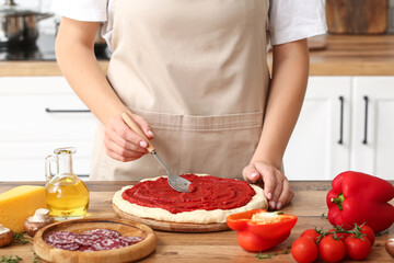 Woman with ingredients spreading tasty tomato sauce on raw pizza dough in kitchen, closeup