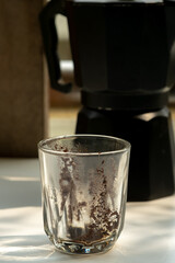 Used glass cup with coffee grounds residue in the foreground, with a blurred black Moka pot and its handle visible in the background, symbolizing a finished morning coffee ritual.