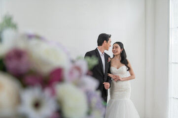 Asian bride and groom in wedding attire smiling and looking at each other, standing indoors by bright window light with floral decoration in foreground. Romantic couple portrait for wedding concept.