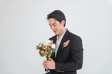 Asian groom in elegant black formal suit holding wedding bouquet, posing indoors with clean white background. Stylish groom portrait for wedding photography concept.