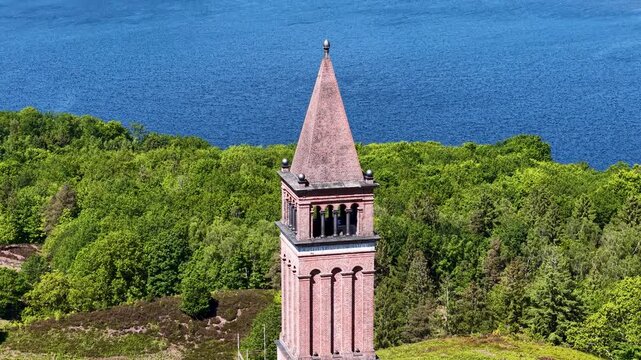 Drone footage showcasing the red-brick Himmelbjerget tower rising above lush green forest and a shimmering lake in Denmark's scenic highlands.