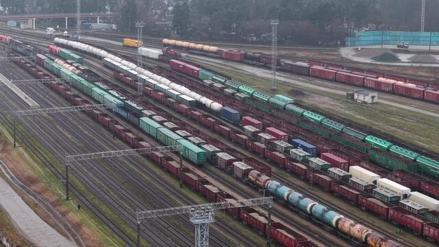 Aerial backward drone shot over multiple cargo tracks in Vilnius, showing long freight trains, tank wagons and colorful containers arranged across parallel rail lines.