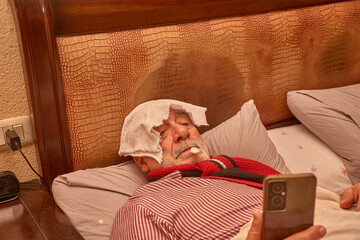 Sick elderly man in bed checking his temperature with a thermometer while looking at his mobile phone in an intimate scene